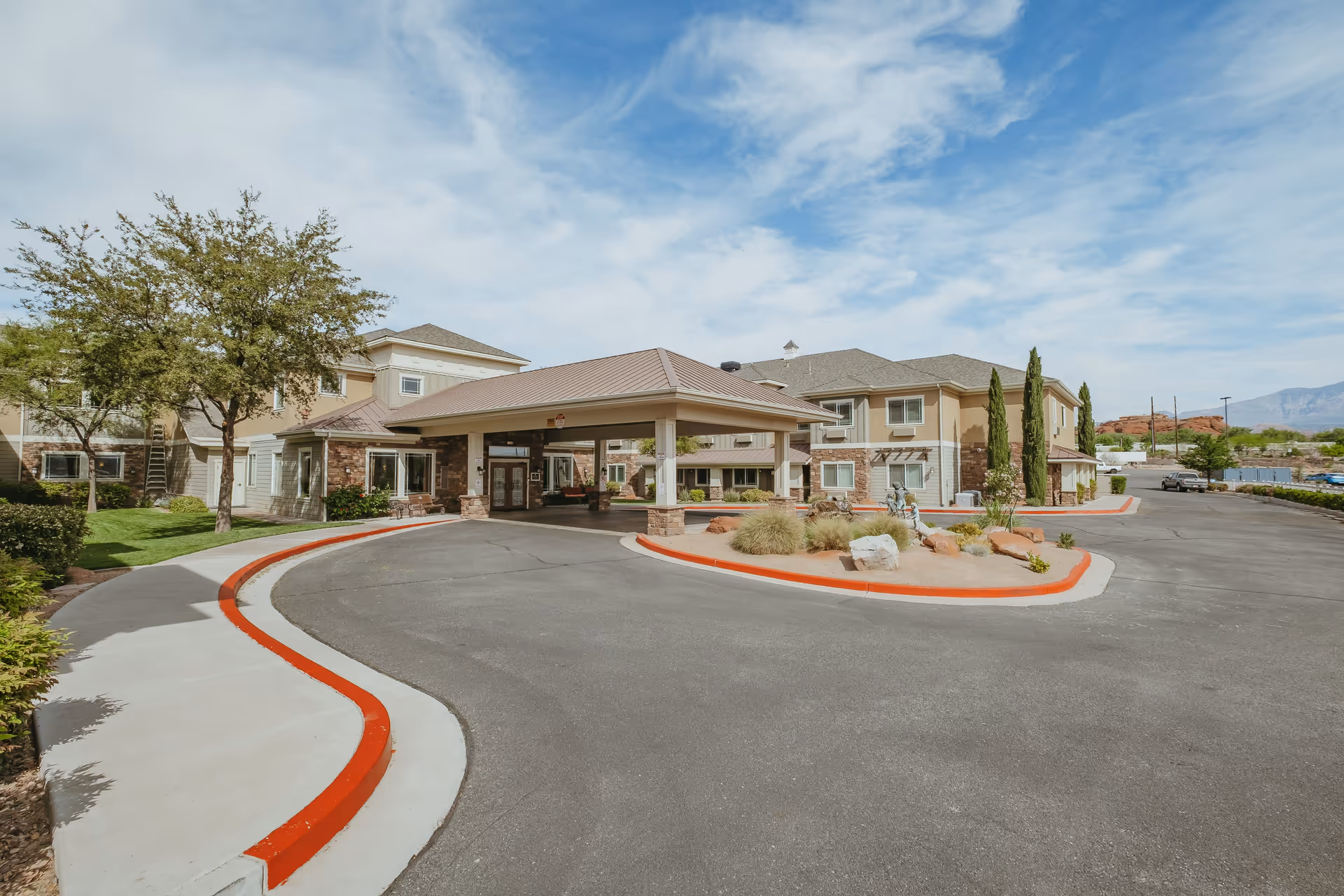 Front entrance and porte-cochere of a senior living facility with a circular drive and landscaped islands under a partly cloudy sky.