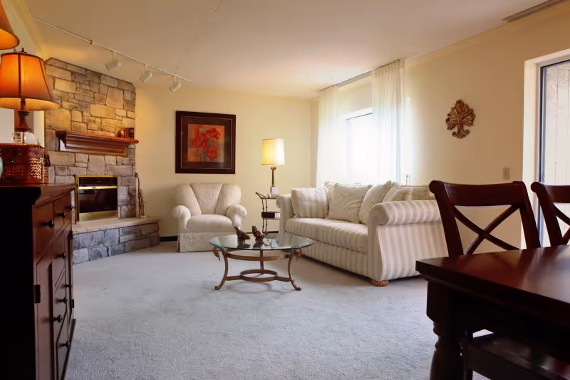 Sunlit living room with a striped sofa, armchair, glass coffee table, stone fireplace, and wooden dining chairs.
