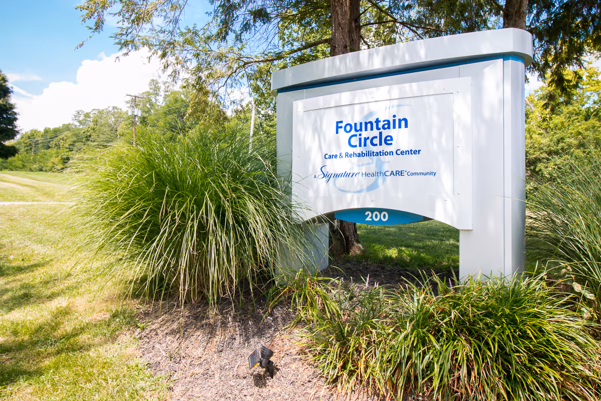 Outdoor view of a white and blue sign for Fountain Circle Care & Rehabilitation Center, surrounded by green grass and plants, with trees and a clear sky in the background.