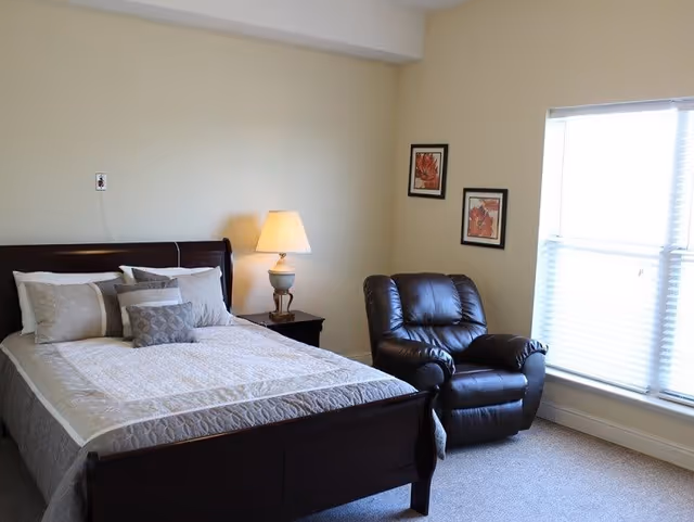 Well-lit bedroom with a made bed, nightstand and lamp, a leather recliner, and a window with blinds.