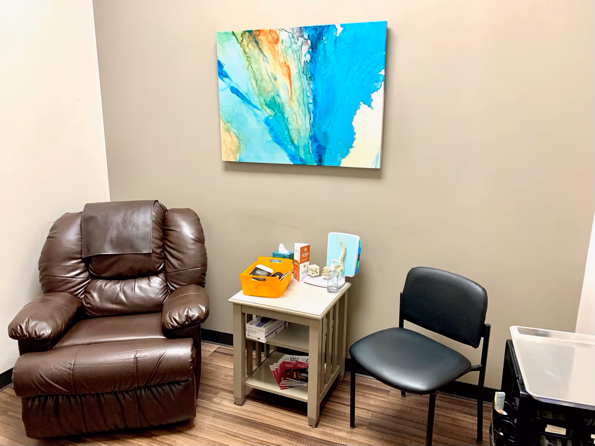 A small infusion center seating area with a brown recliner, black chair, and abstract wall art.