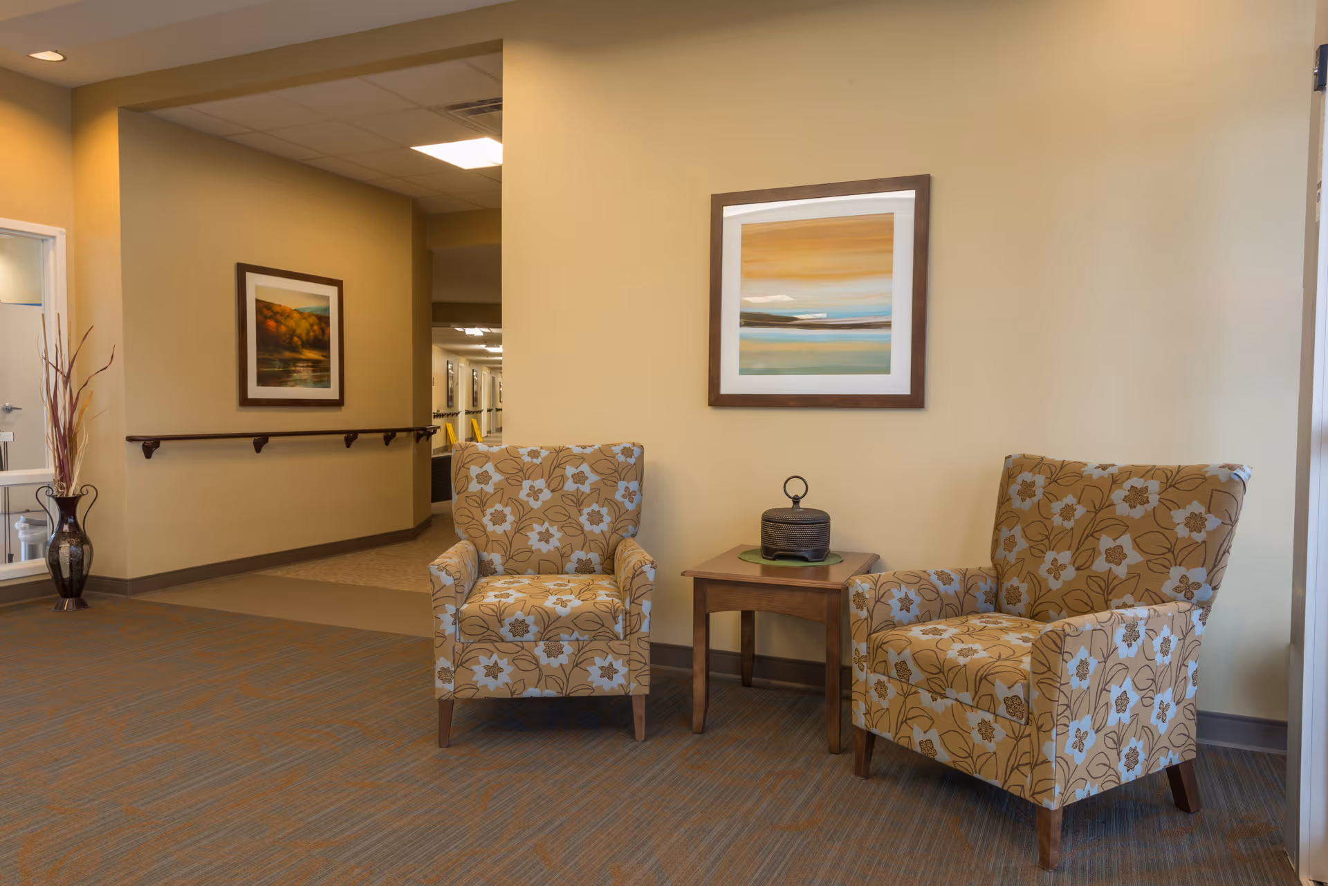 Two patterned armchairs flank a small table in a beige senior living lobby, artwork on wall.