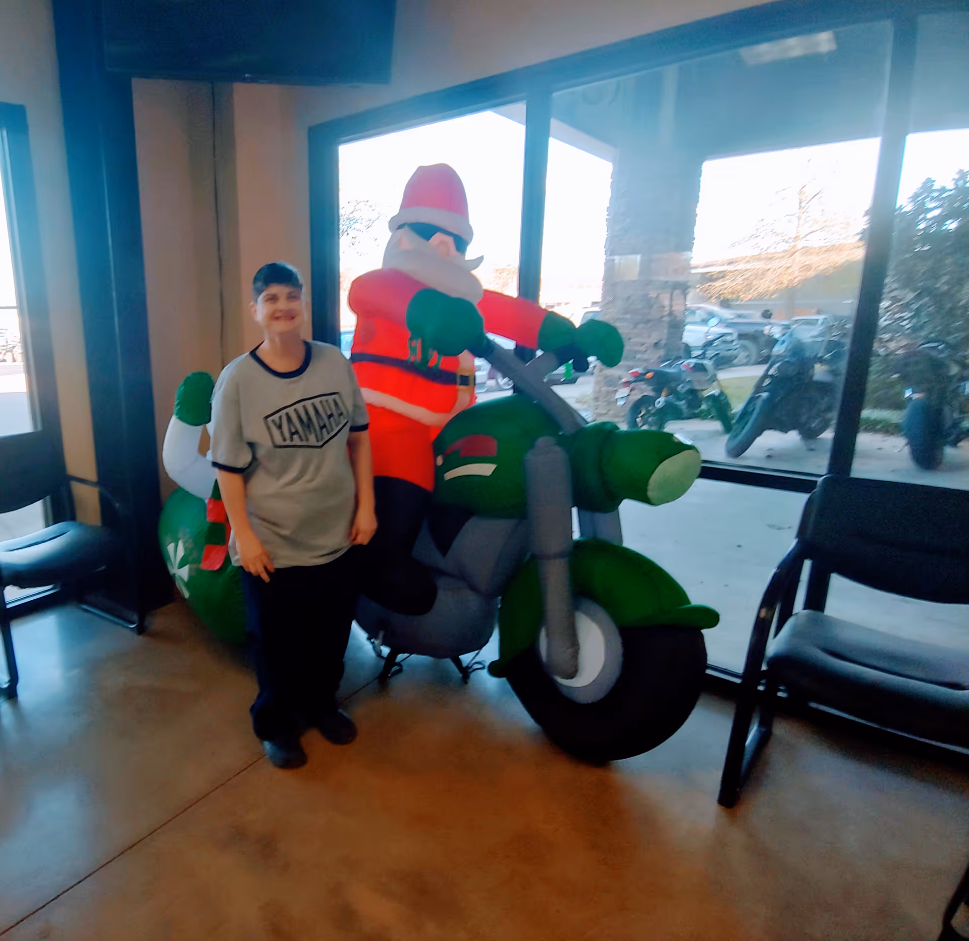 A person stands beside a large inflatable Santa on a green motorcycle indoors near windows.