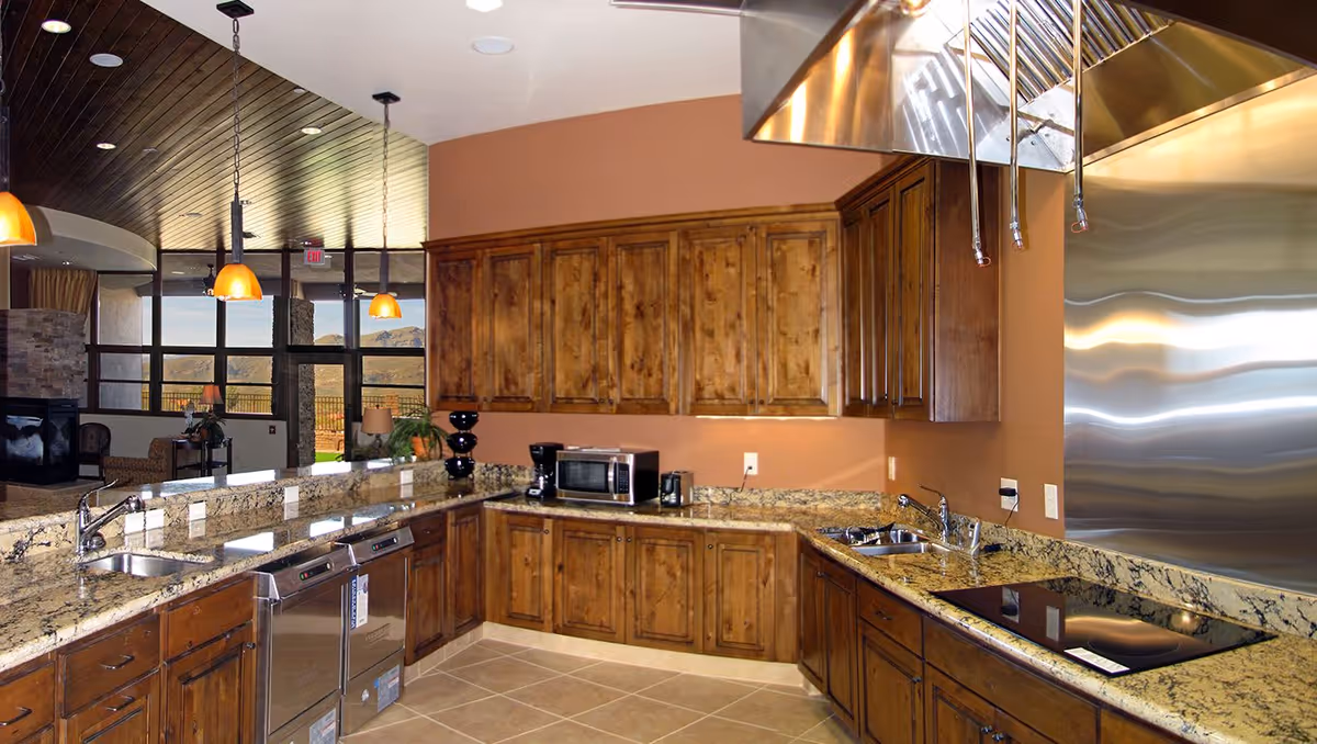 A spacious kitchen with wooden cabinets, granite countertops, two sinks, a microwave, a coffee maker, a dishwasher, and a stainless steel range hood. The kitchen opens into a living area with large windows showing a scenic mountain view outside.