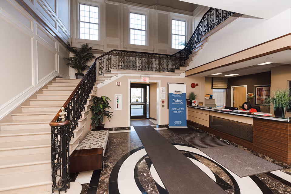 Interior view of the lobby at Glengariff Rehabilitation and Healthcare Center featuring a grand staircase with ornate black railings, a reception desk with a staff member, potted plants, and large windows letting in natural light.