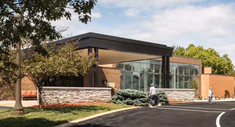 Front entrance of a brick senior living building with a covered porte-cochère, landscaped beds, and two people walking near the driveway.