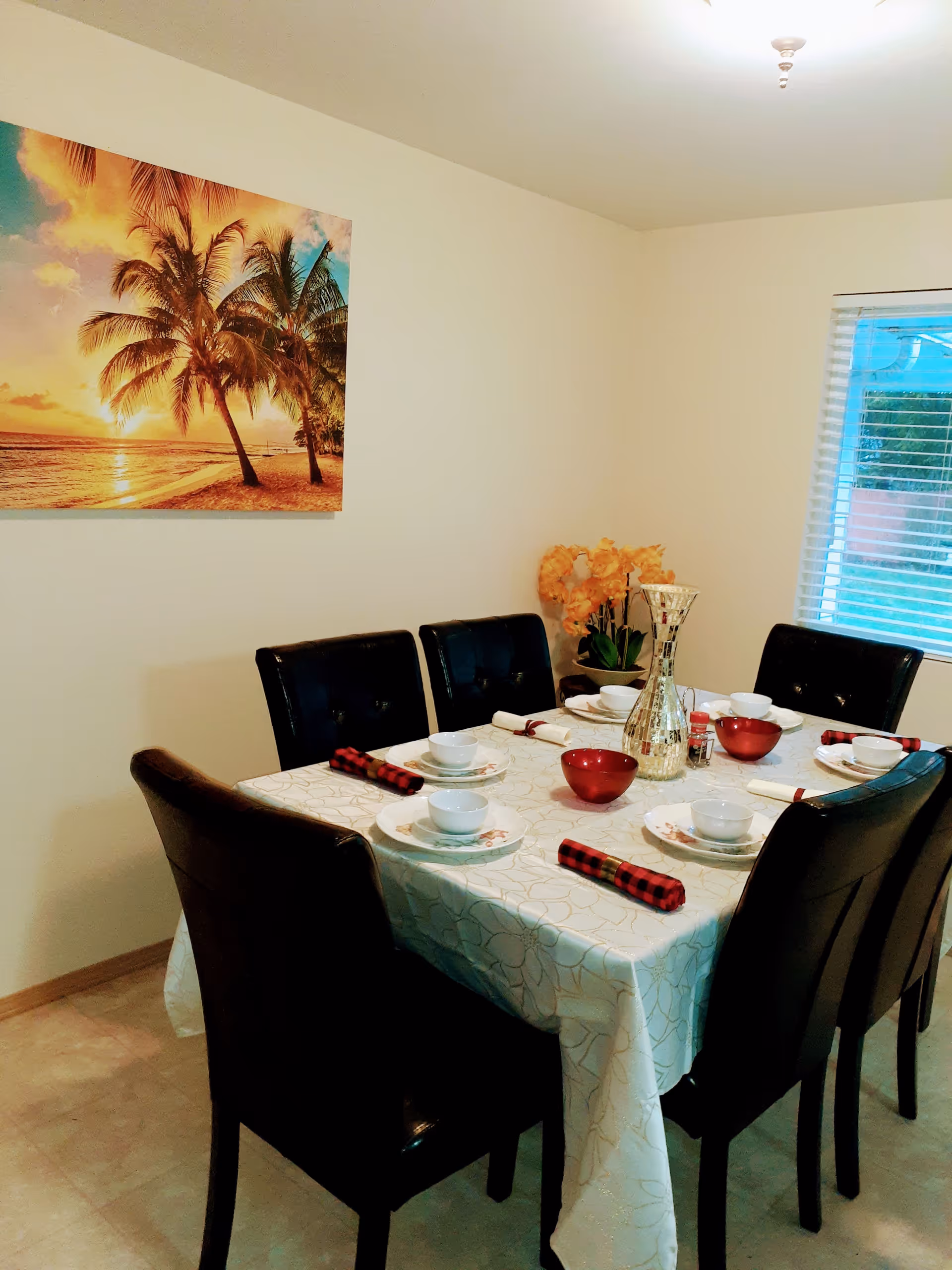 A dining room with a rectangular table covered with a white patterned tablecloth. The table is set with white plates, bowls, red and black checkered napkins, and red bowls. Six black leather chairs surround the table. On the wall is a large picture of a beach sunset with palm trees. A window with blinds is on the right side, and a vase with orange flowers is placed on a small table in the corner.
