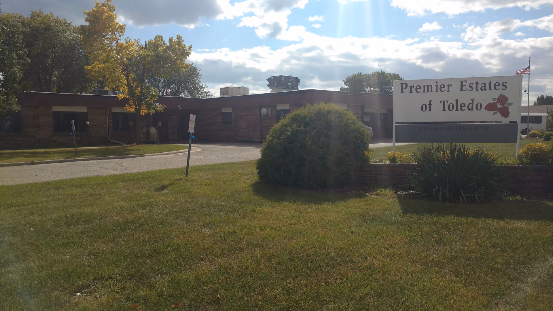 Exterior view of a single-story brick building with a lawn and trees under a partly cloudy sky. A sign in front reads 'Premier Estates of Toledo' with a flower graphic and an American flag is visible in the background.