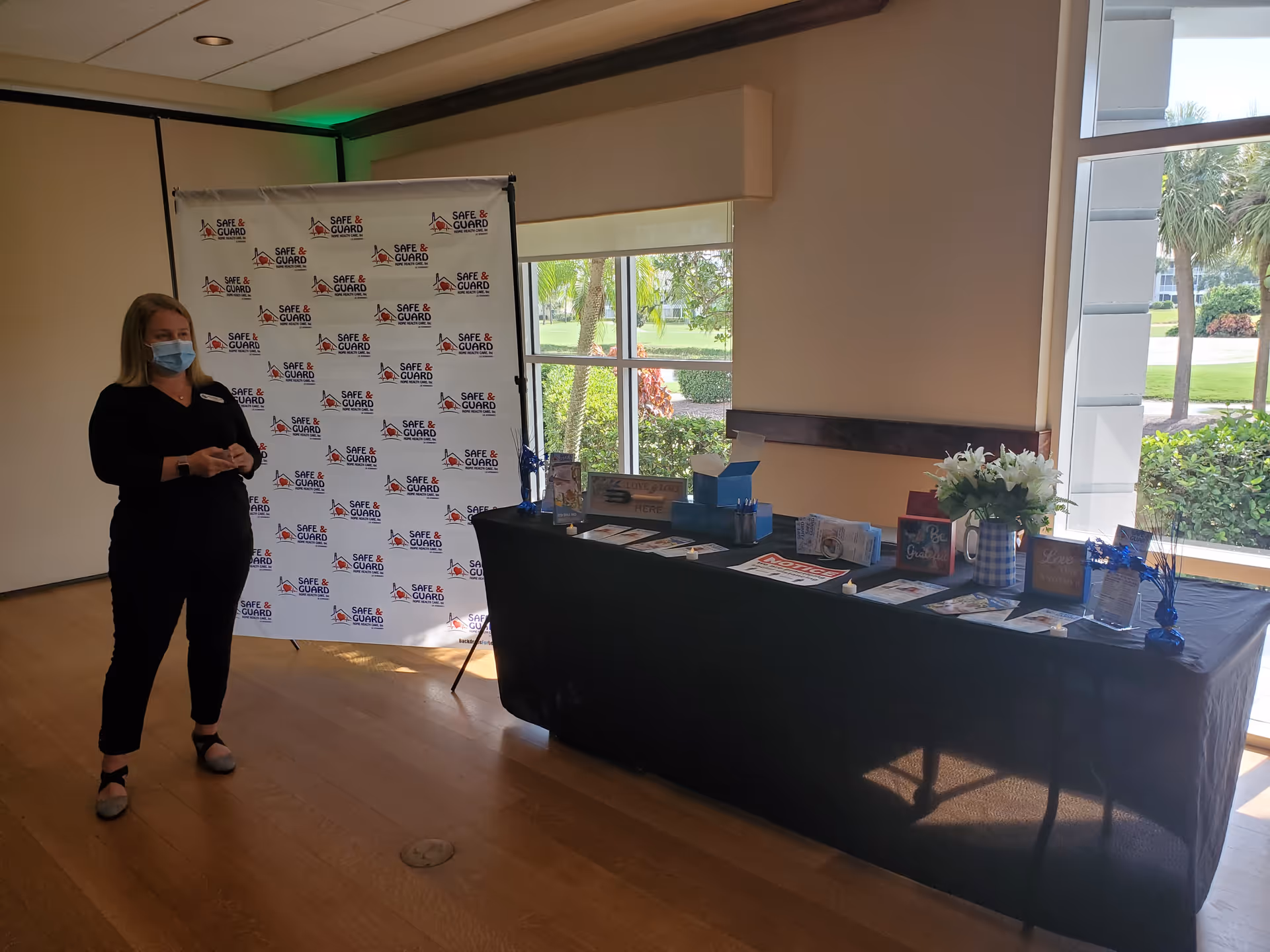 A masked staff member stands beside a branded banner and table with brochures indoors.