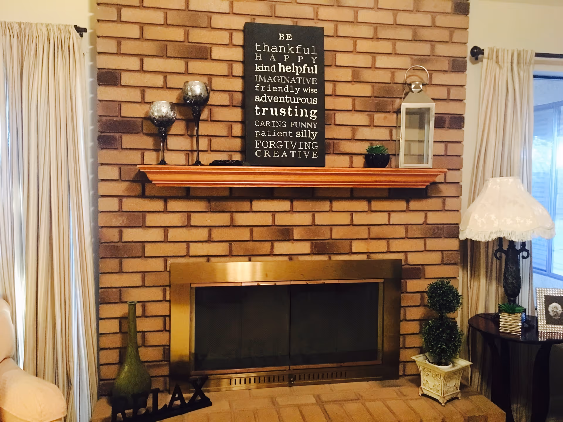 A cozy living room corner featuring a brick fireplace with a brass frame. Above the fireplace is a wooden mantel decorated with two silver candle holders, a small potted plant, a lantern, and a black sign with inspirational words. On the hearth, there is a green vase and a decorative 'RELAX' sign. To the right, a small round table holds a lamp with a white lace shade and a framed photo. Cream-colored curtains frame the windows on either side.
