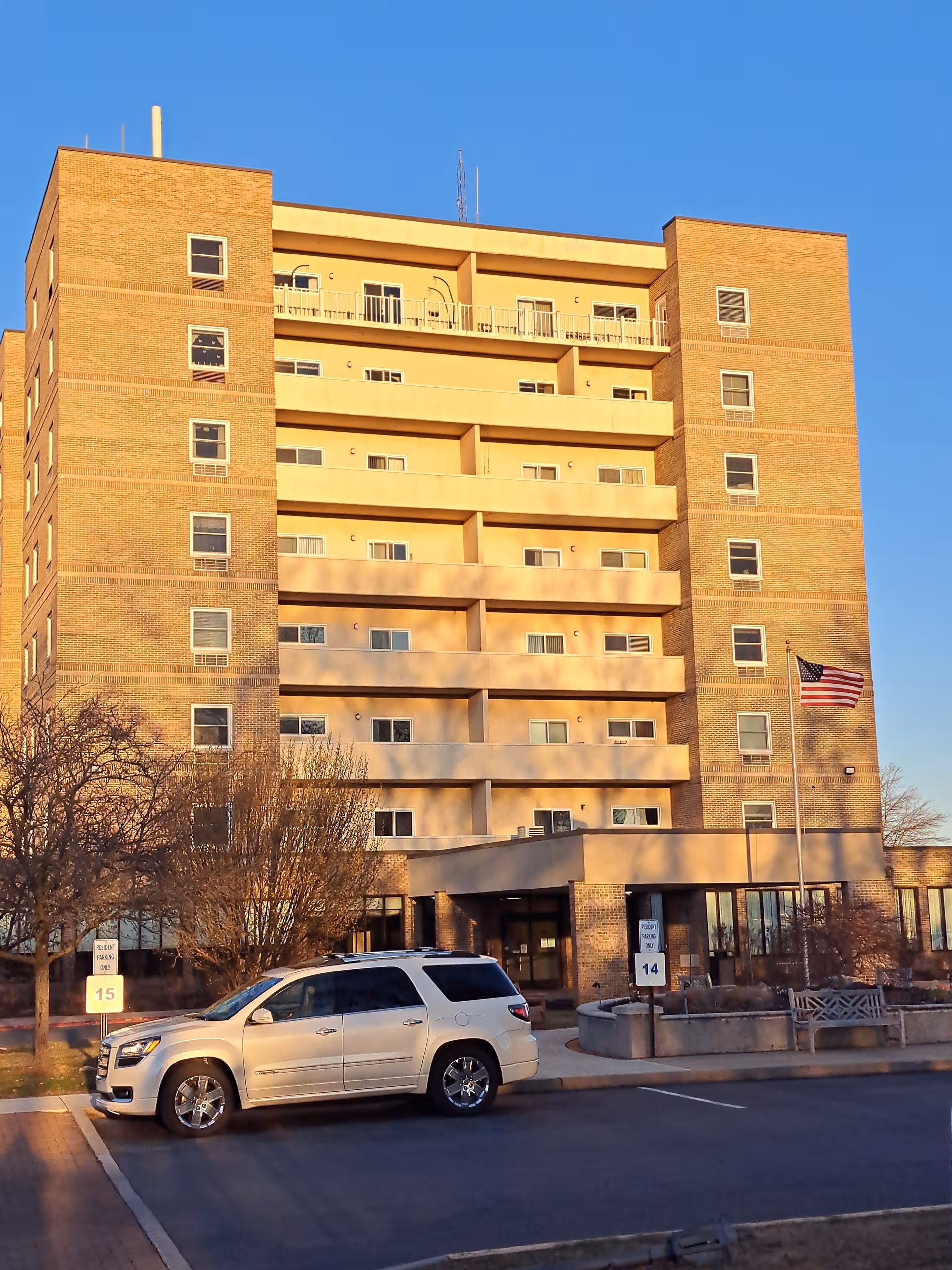 Six-story brick senior living building with entrance, flag, and a white SUV parked outside.
