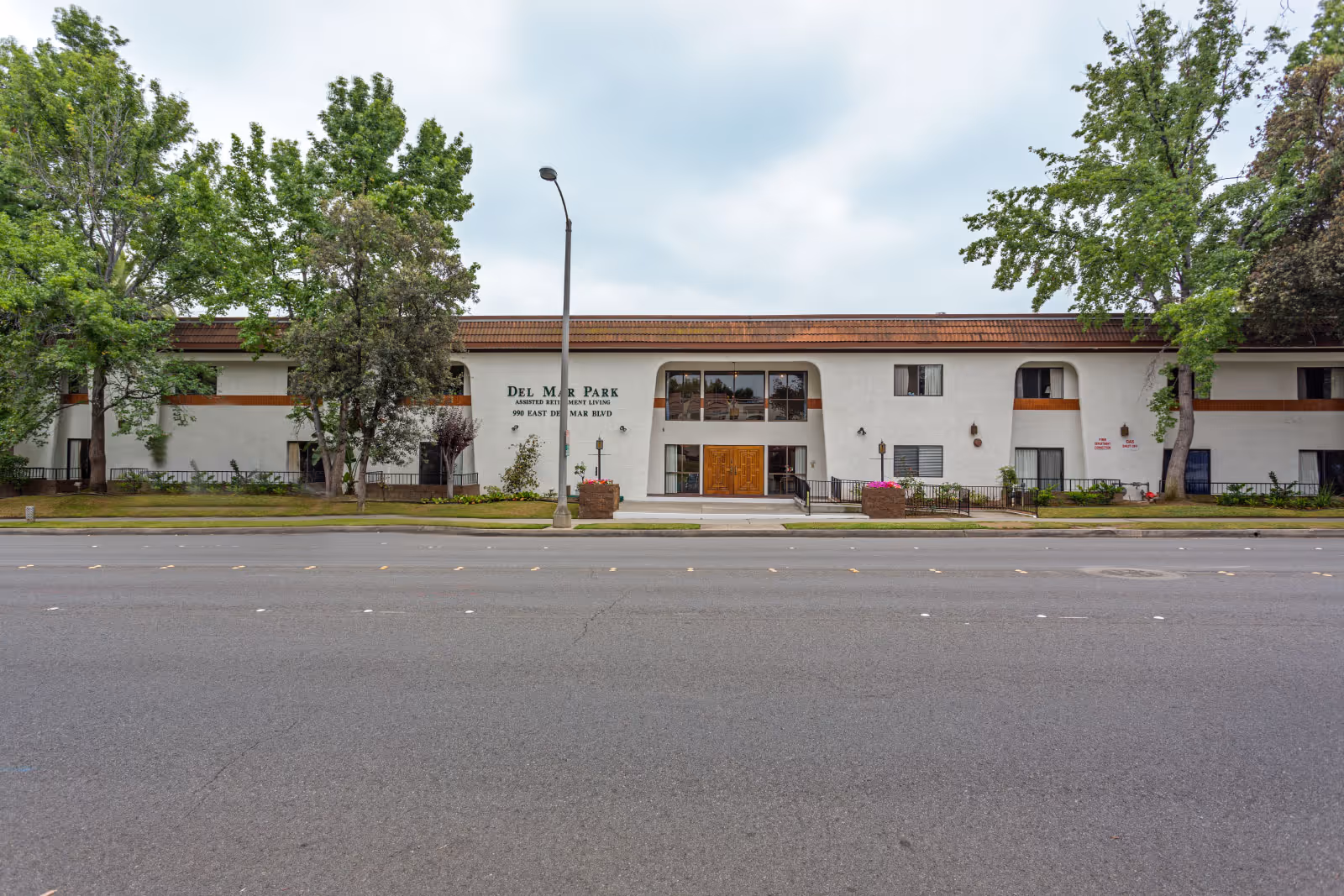 Front exterior view of Del Mar Park assisted retirement living facility, a two-story white building with brown roof trim, surrounded by trees and a sidewalk in front.
