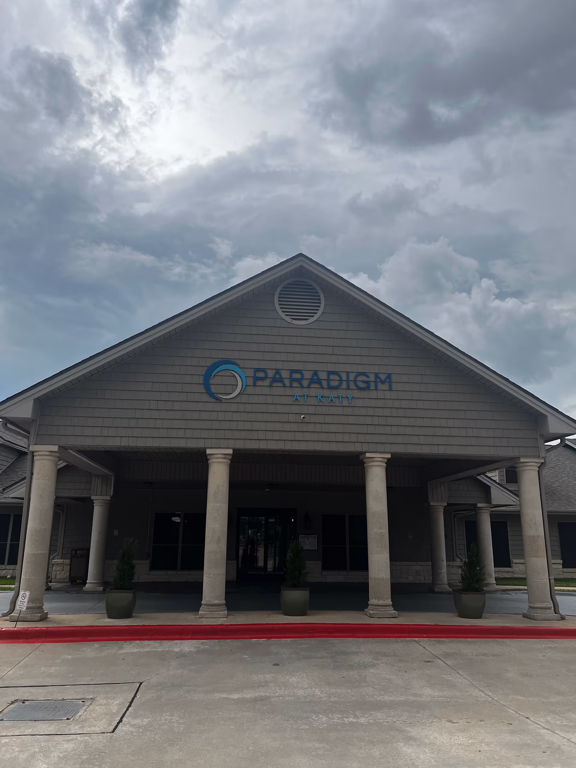 Front view of a beige, column-supported building with a blue Paradigm at Katy sign and entrance.
