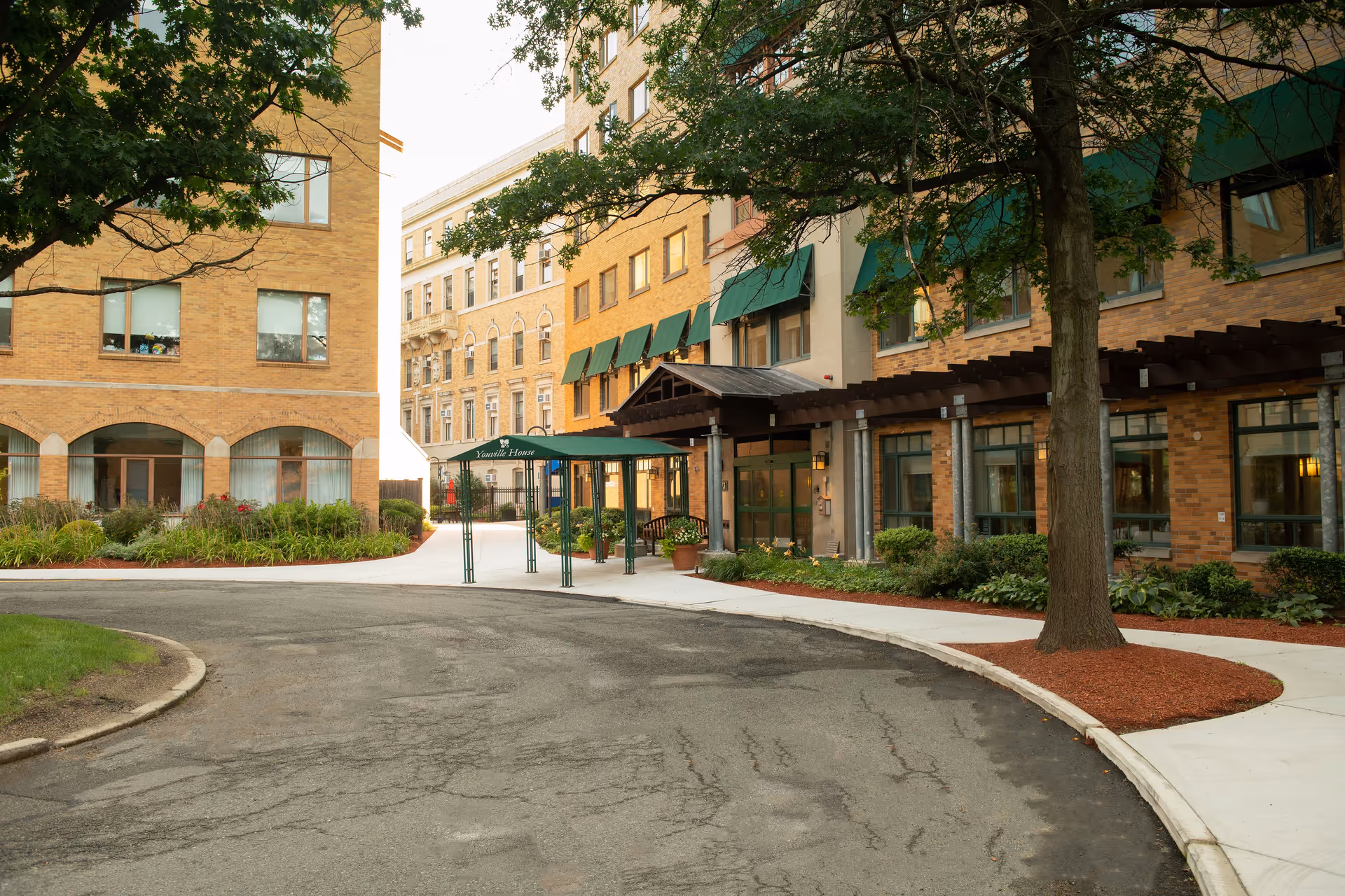 Front entrance of Youville House Assisted Living, showing the brick facade, green awnings, covered drop-off canopy, trees and landscaping.