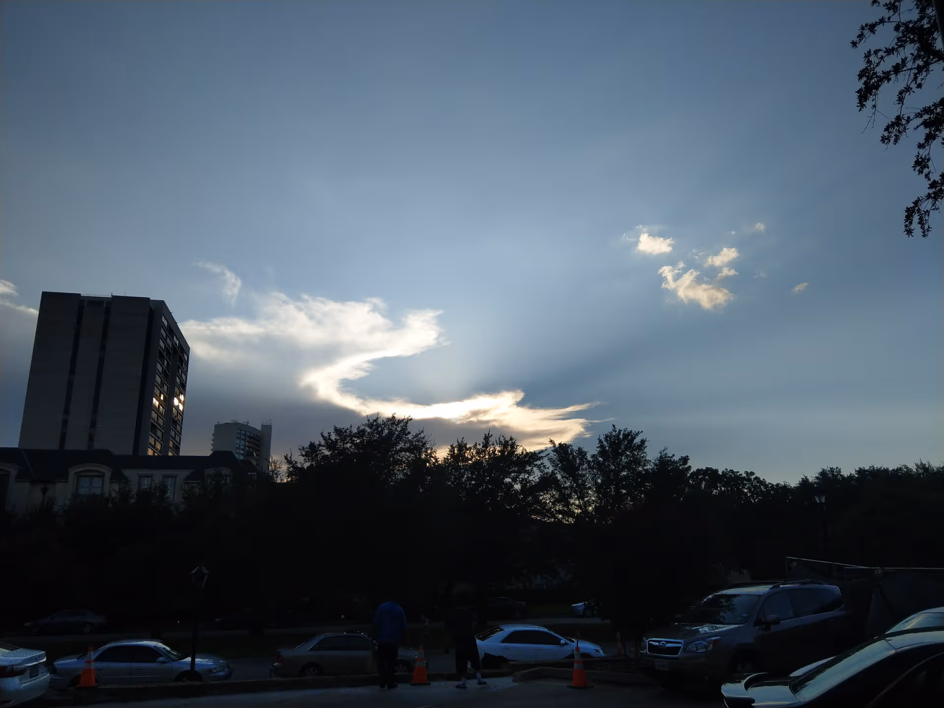 Dusk exterior view with sun rays breaking through clouds behind a tall building and trees