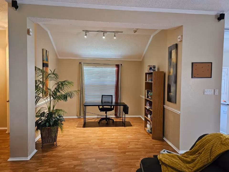 Interior study area with desk, chair, bookshelf, plant, and window blinds.
