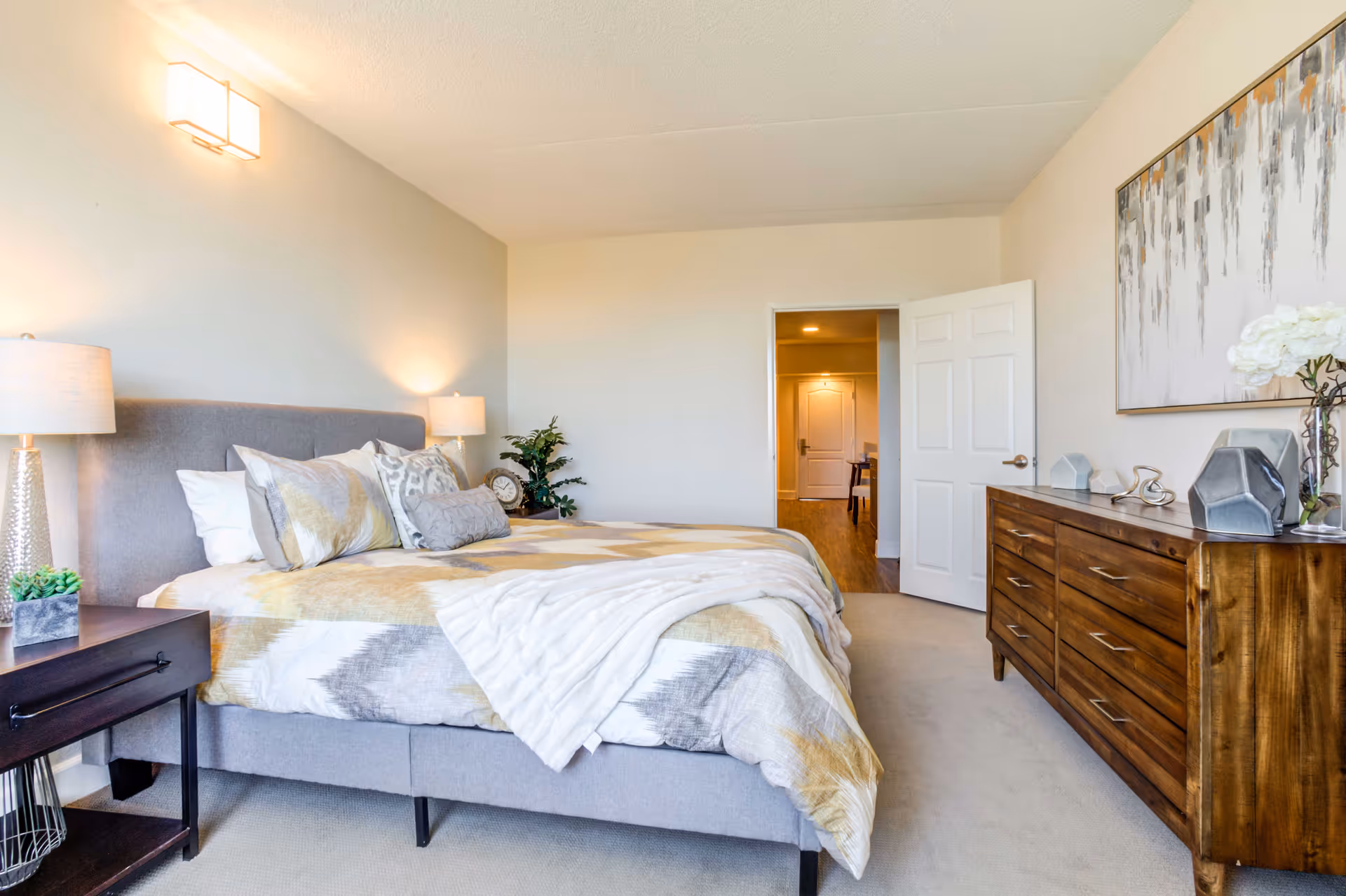 A bright and neatly arranged bedroom featuring a large bed with patterned bedding and multiple pillows. Two bedside tables with lamps flank the bed. A wooden dresser with decorative items and a large abstract painting hangs on the wall. The door is open, showing a hallway with wooden flooring and a closed door at the end.