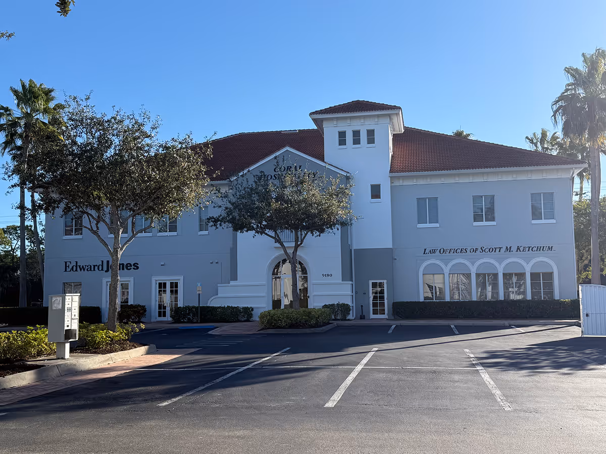 White two-story building with a red-tiled roof and arched windows in a parking lot.