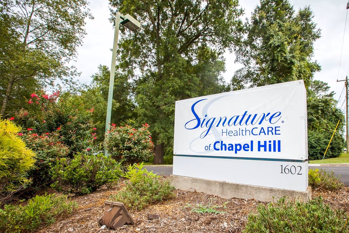Outdoor view of a sign for Signature HealthCARE of Chapel Hill surrounded by greenery and trees, with a street and utility poles in the background.