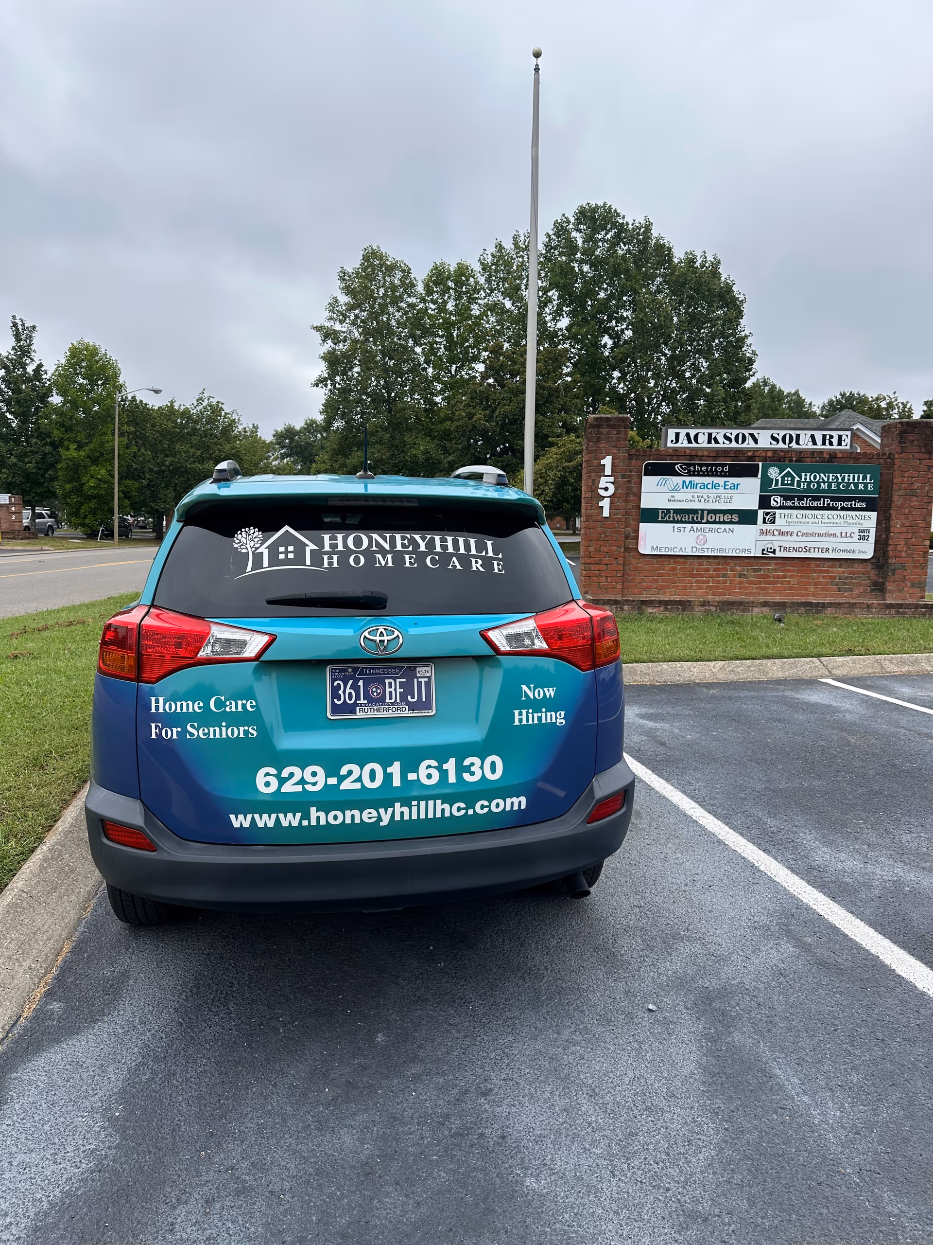 Back of a blue HoneyHill HomeCare van parked in a lot beside a sign.