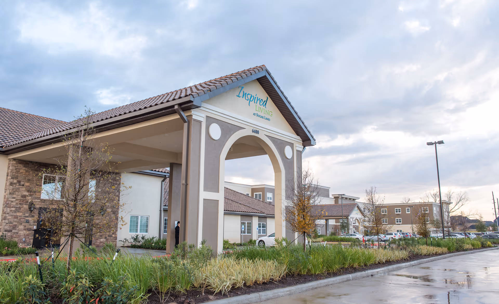 Entrance porte-cochère and facade of a senior living building with landscaped grounds and parked cars under a cloudy sky.