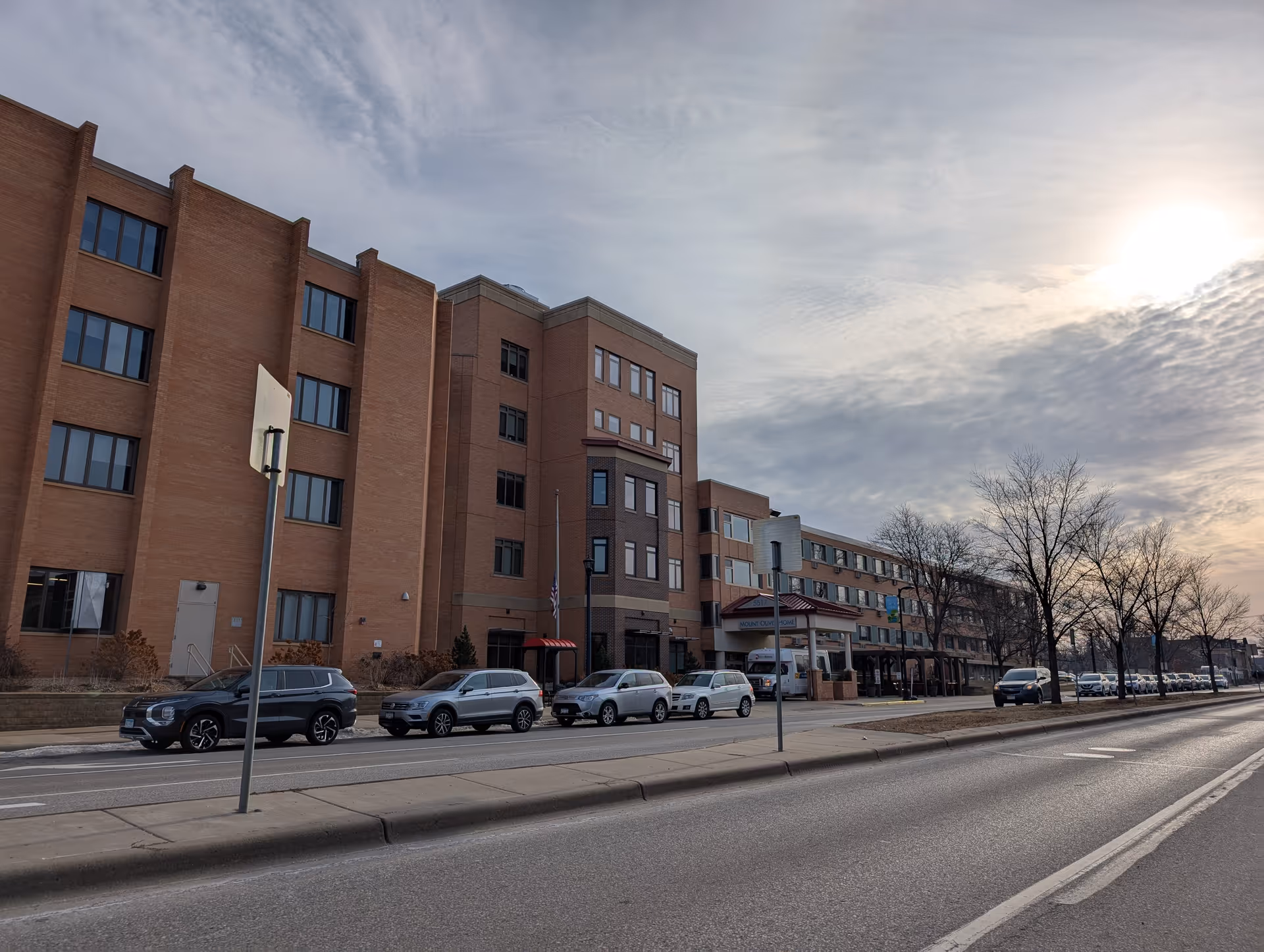 Street view of a multi-story brick care facility with cars parked along the curb under a cloudy sky.