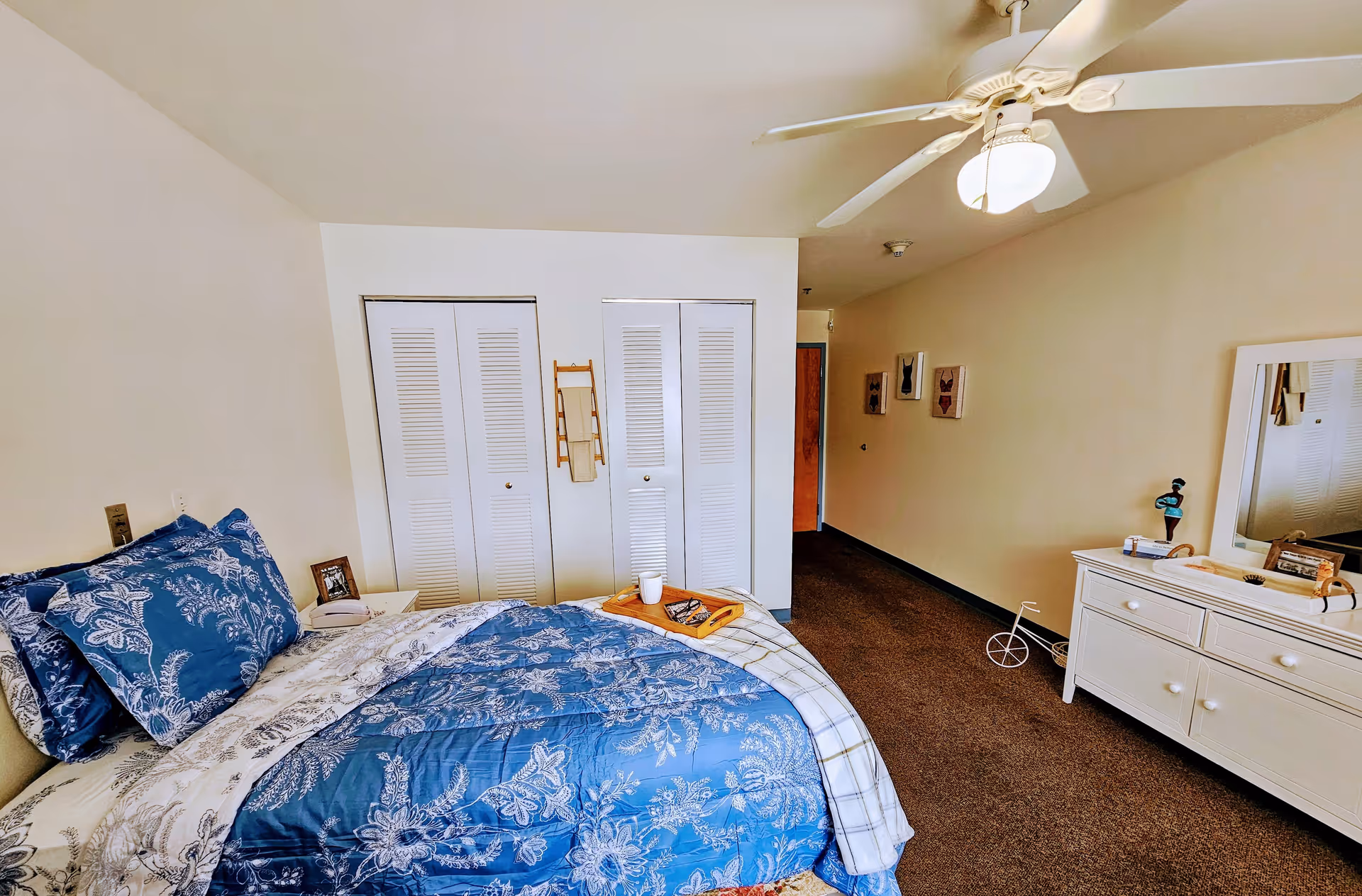 Bedroom with blue floral bedding, ceiling fan, white dresser and mirror, and double closet doors.