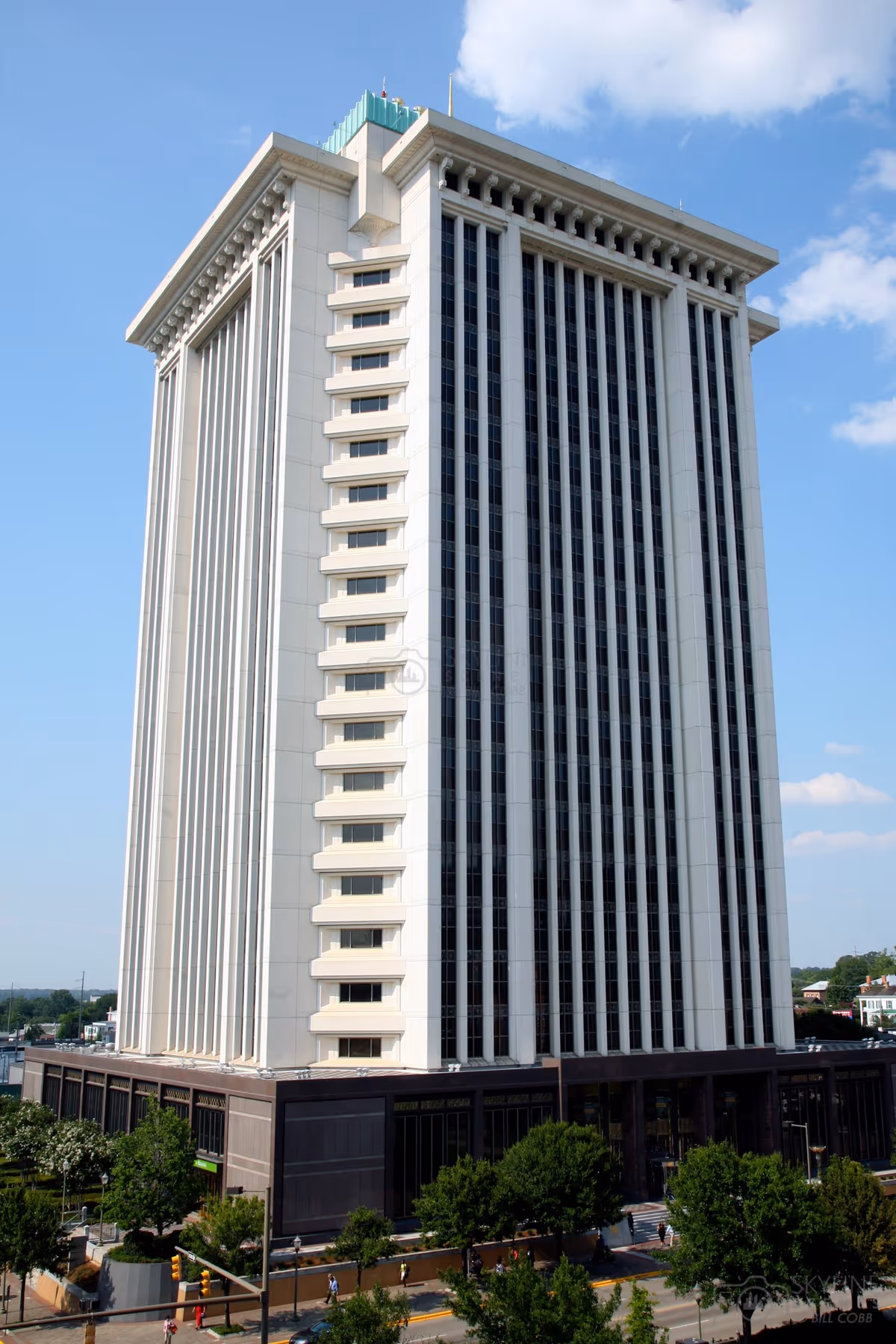 Tall white high-rise building with vertical windows dominates the skyline, street and trees in foreground.