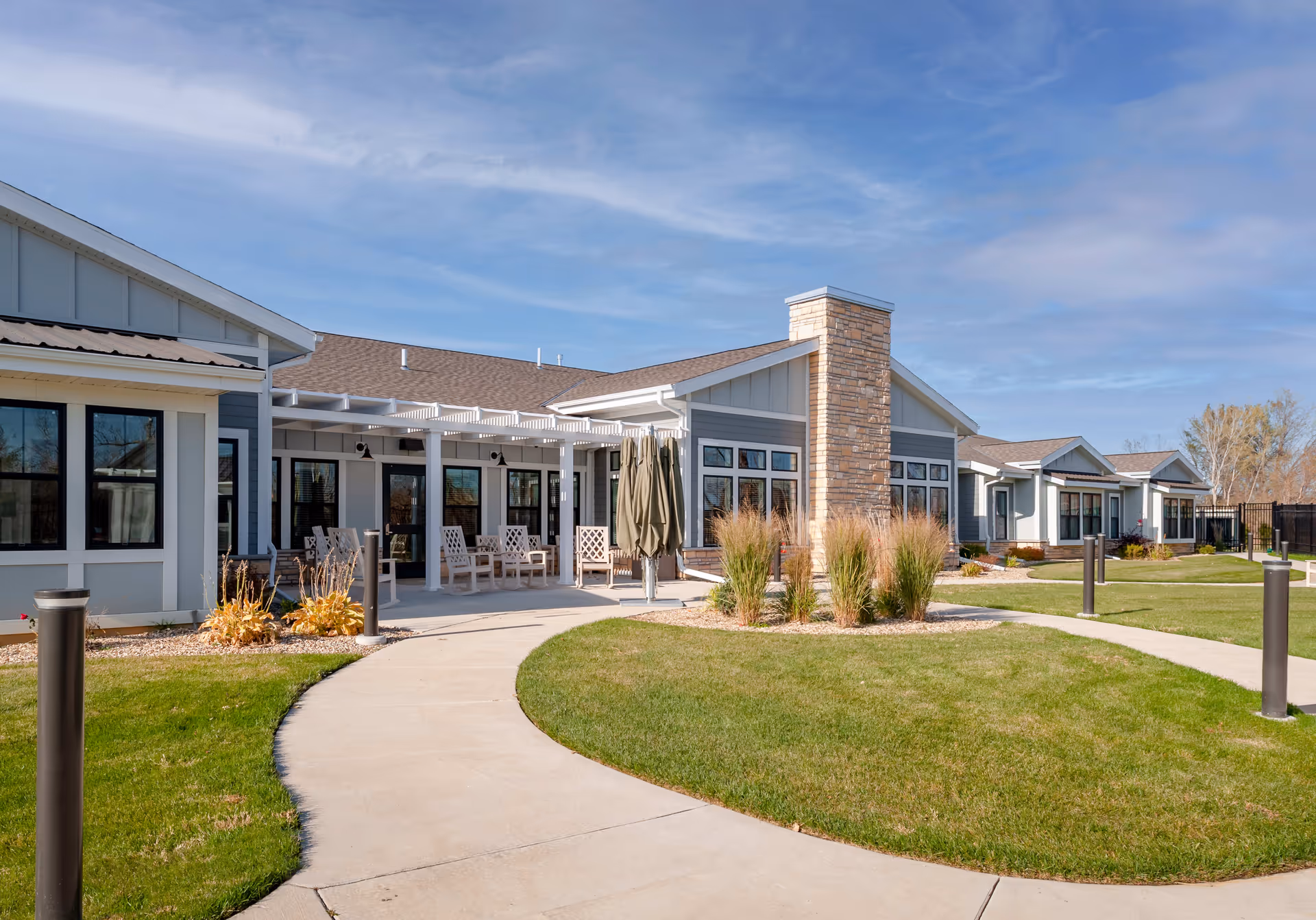 Exterior view of a single-story senior living building with a covered patio, seating, curved walkway and landscaped lawn under a blue sky.