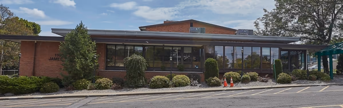 Front exterior of a low brick building with large windows, shrubs, and a covered entrance walkway.