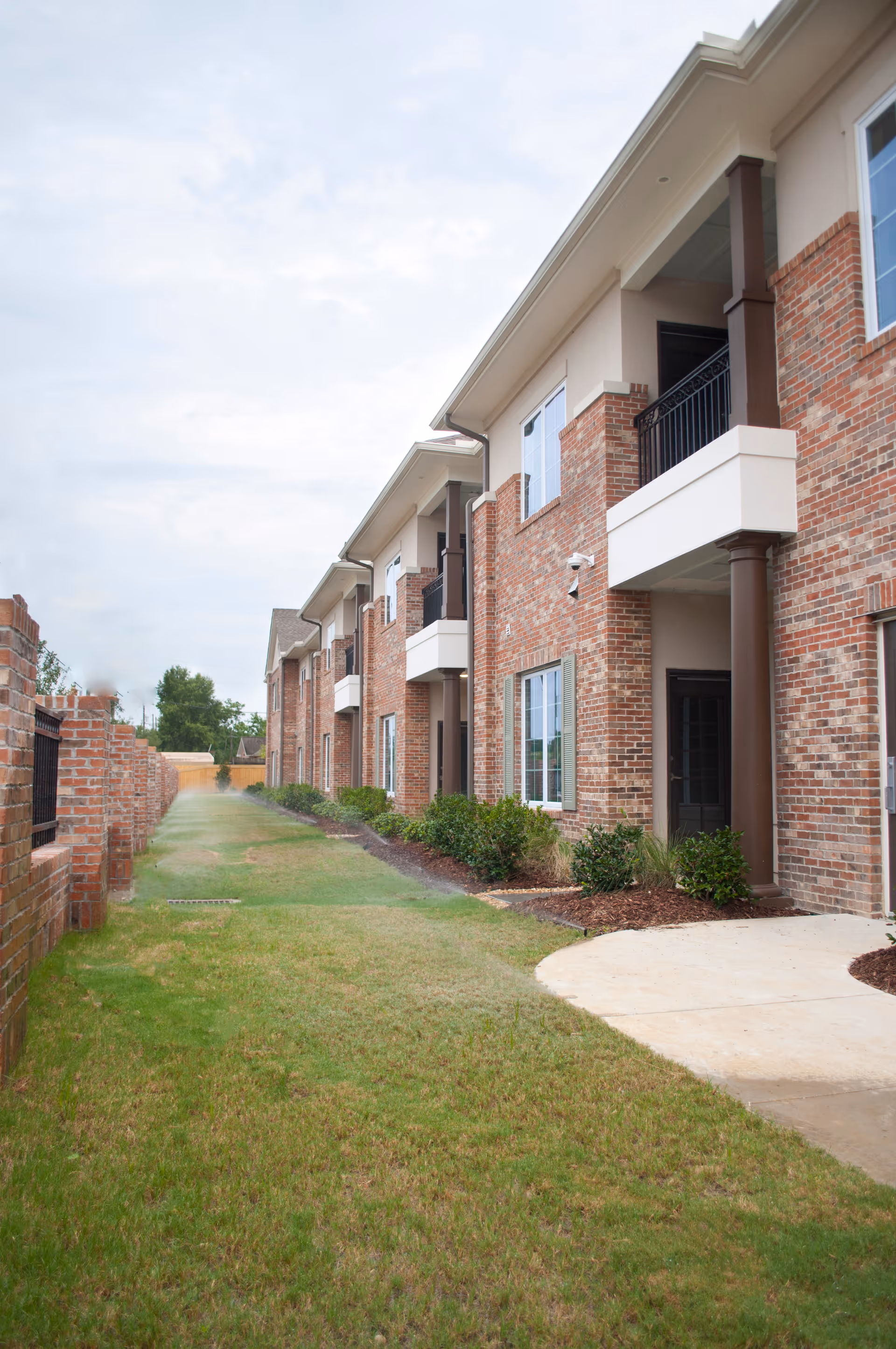Two-story brick building with balconies, windows, and a grassy walkway along the facade.
