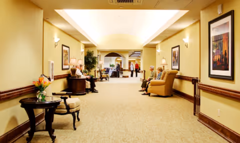 Well-lit, carpeted common corridor in an assisted living facility with armchairs, side tables, framed art, and several residents seated and standing.