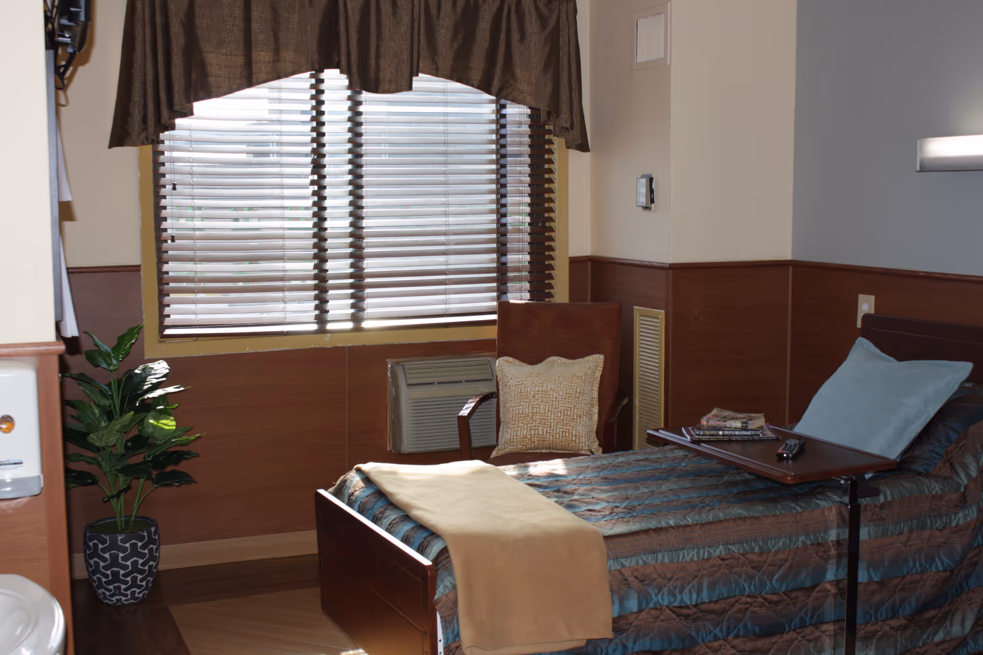 A cozy bedroom in Highland Care Center featuring a single bed with a blue and brown striped comforter, a beige blanket, and a blue pillow. Next to the bed is a brown chair with a decorative cushion, a small table with magazines and a remote control, a window with blinds and brown curtains, and a potted plant in the corner. The room has wood paneling on the lower half of the walls and a light fixture above the bed.