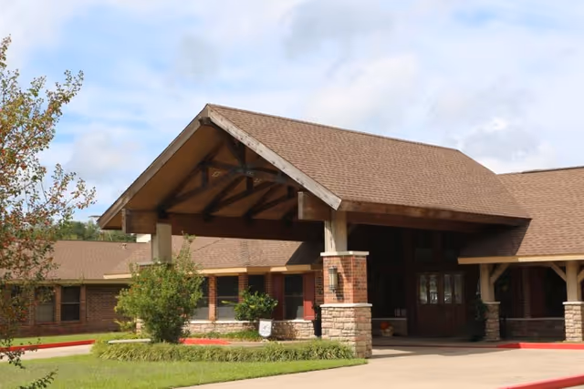 Exterior view of Timberwood Nursing & Rehabilitation Center showing the main entrance with a large covered driveway supported by stone and wooden pillars, surrounded by green landscaping and a partly cloudy sky.