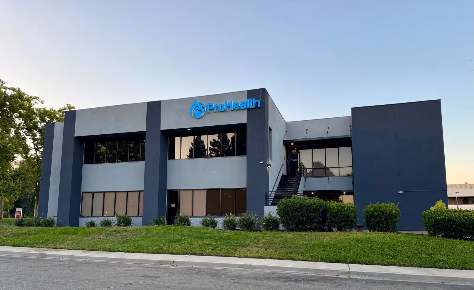 Modern two-story ProHealth building with blue accents, large windows, and neatly landscaped front lawn.