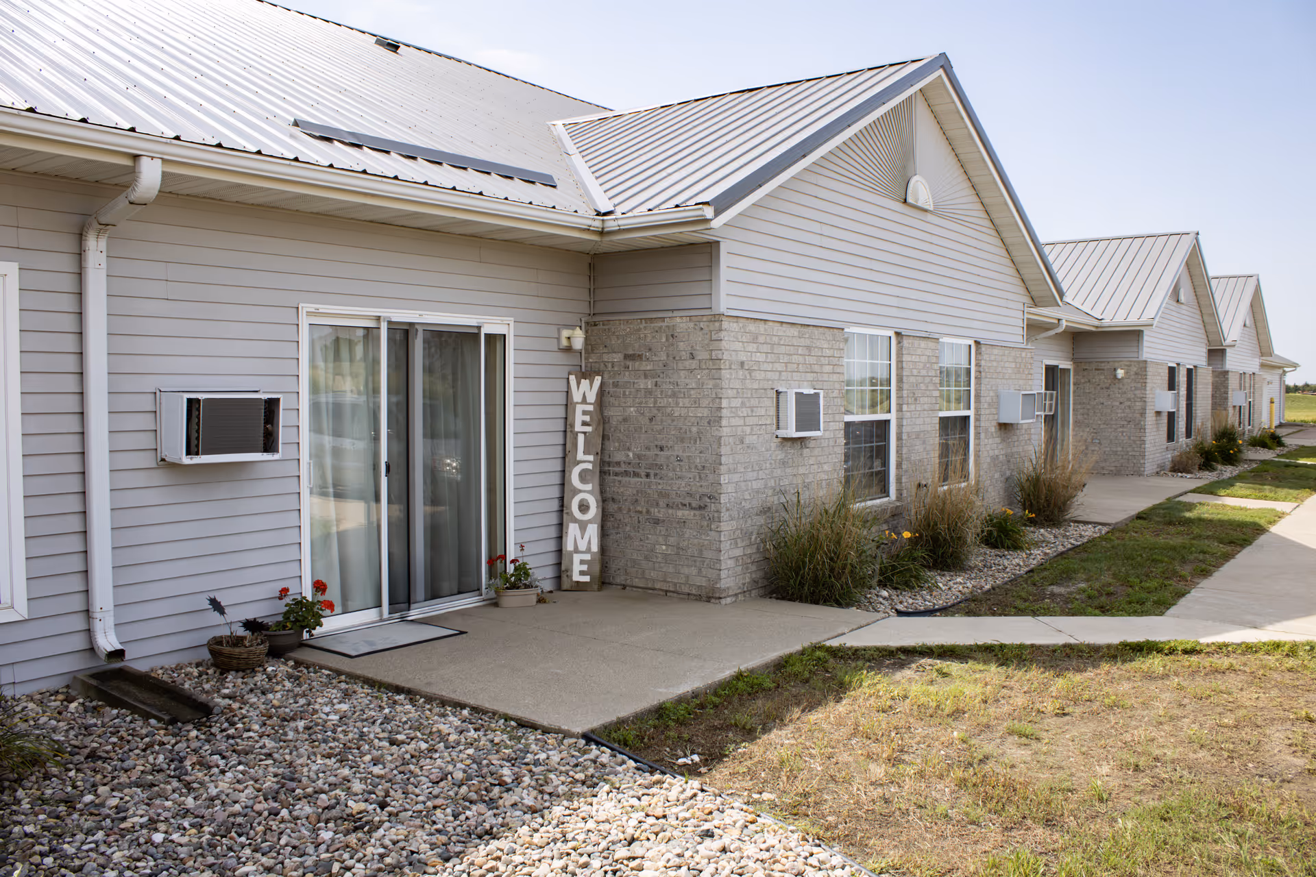 Row of light gray senior living cottages with metal roofs and a welcome sign.