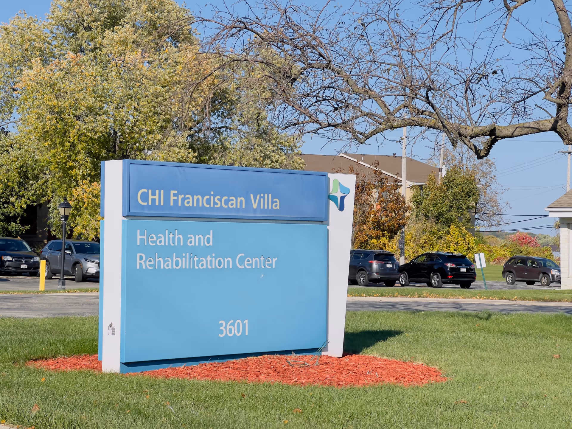 A blue outdoor sign reading 'CHI Franciscan Villa Health and Rehabilitation Center 3601' on a grassy lawn with parked cars and trees behind.