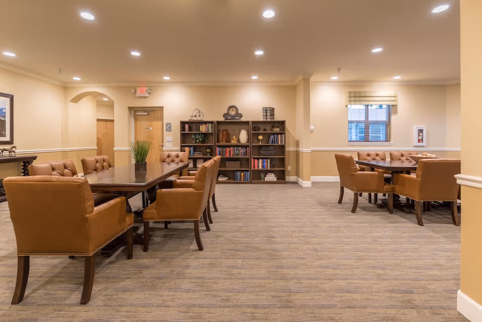 Warm senior living common area with brown upholstered chairs around tables and a bookshelf.
