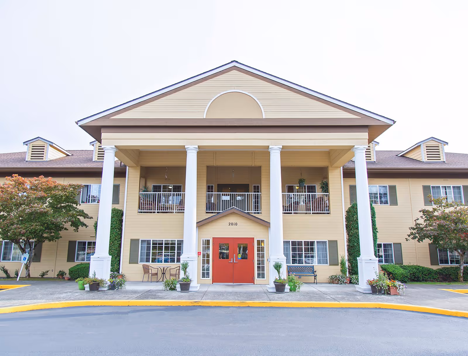Front entrance of a two-story beige assisted living building with tall white columns, red double doors, balconies, and landscaping.