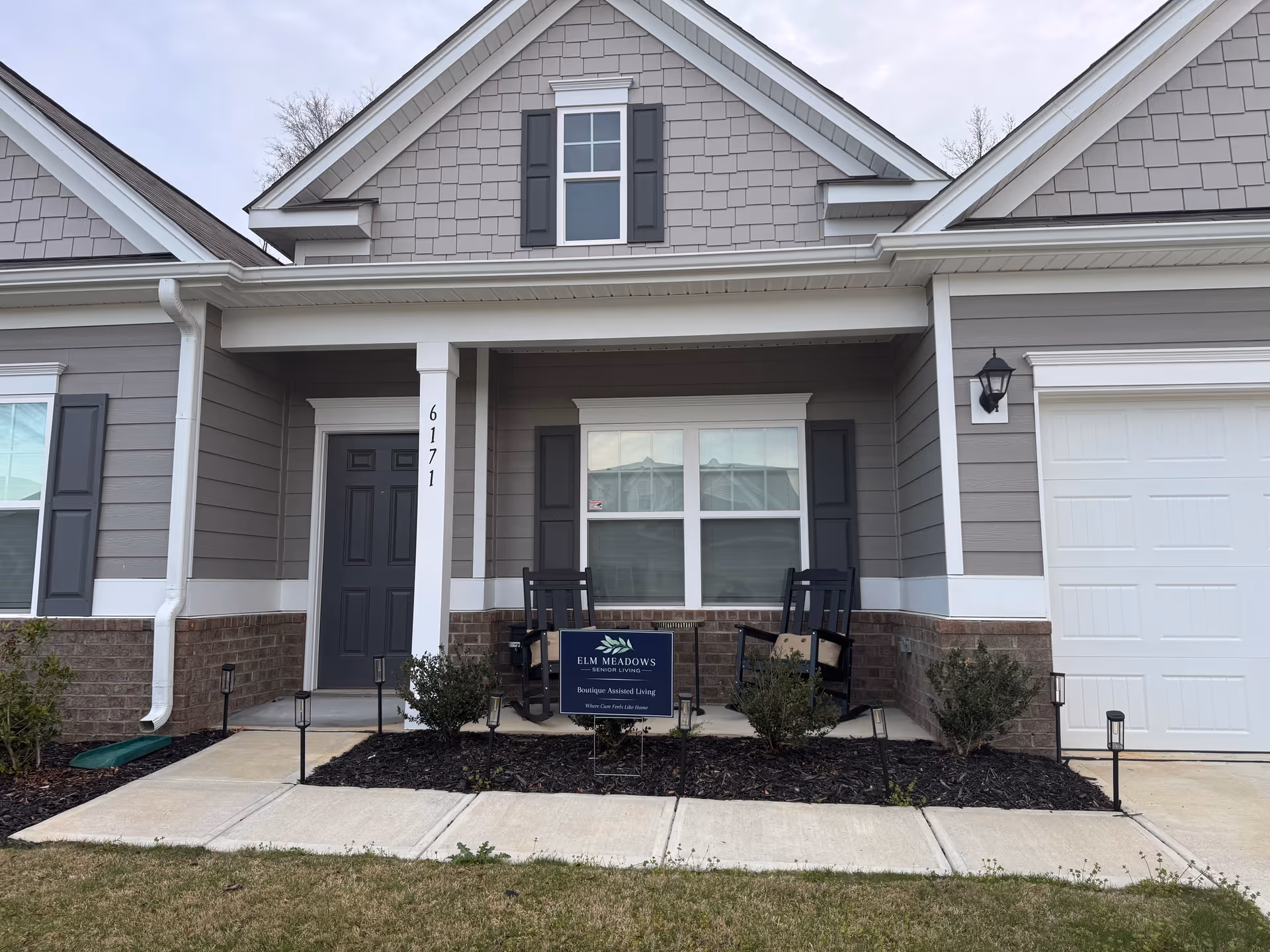 Gray, two story senior living facility exterior with front porch, windows, and rocking chairs.