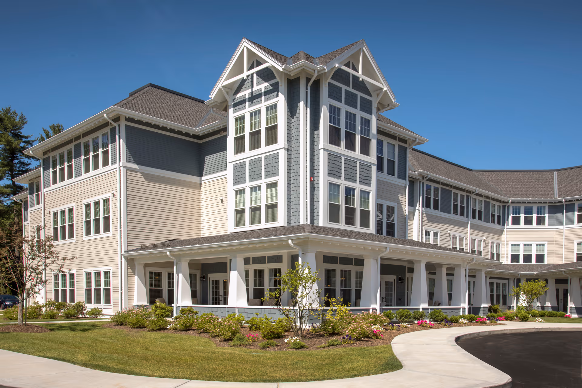 Three-story senior living facility building with a wraparound porch, landscaped flower beds and a curved driveway under a clear blue sky.