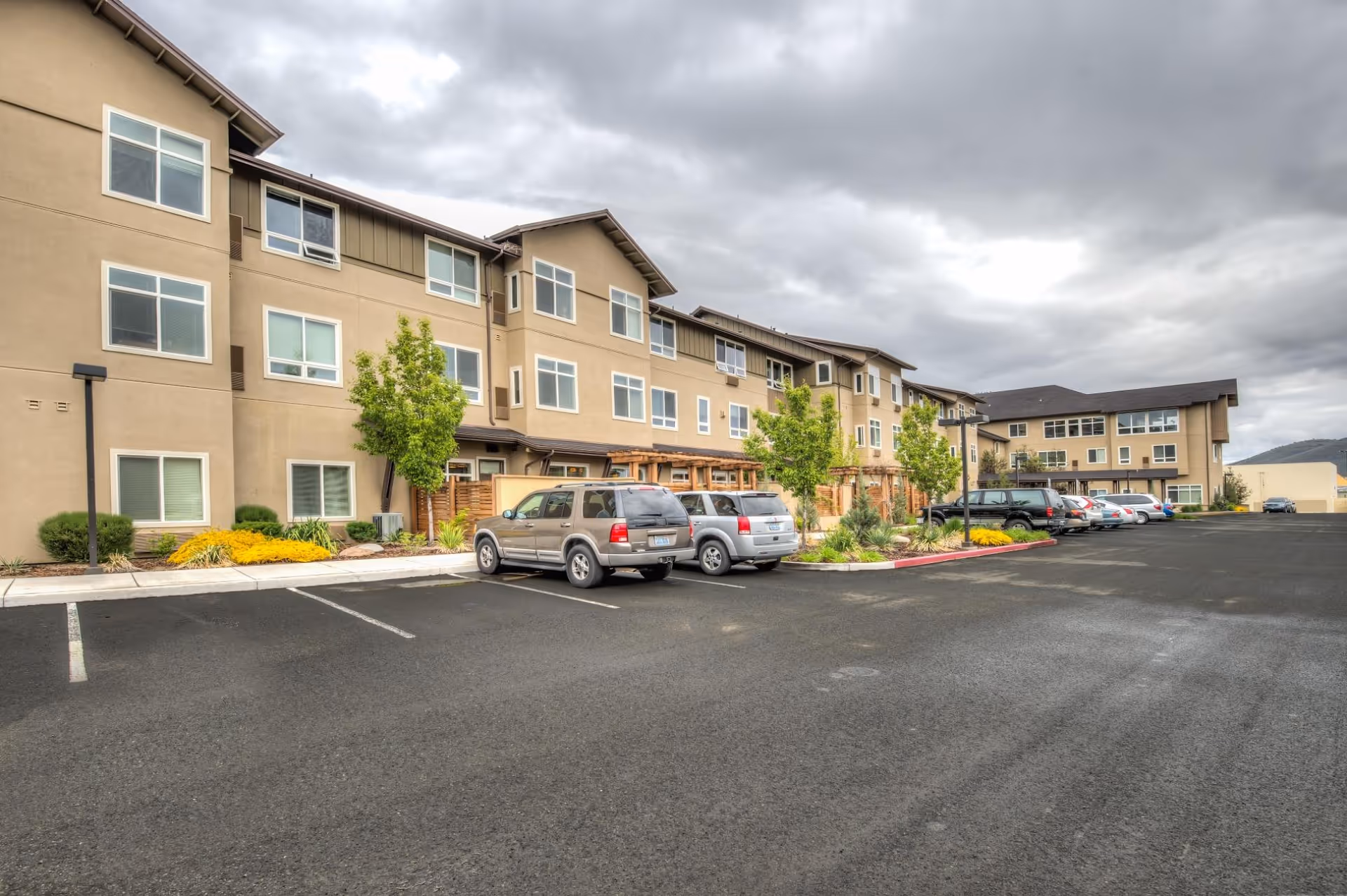 Exterior view of a three-story assisted living facility with beige walls and multiple windows. Several cars are parked in the parking lot in front of the building. There are small trees and landscaped areas along the building. The sky is cloudy.