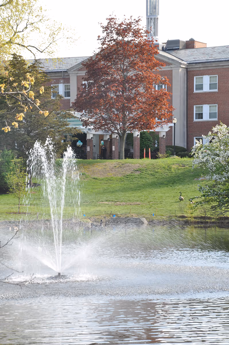 A brick three-story building with a tree-lined entrance overlooks a pond with a central fountain.
