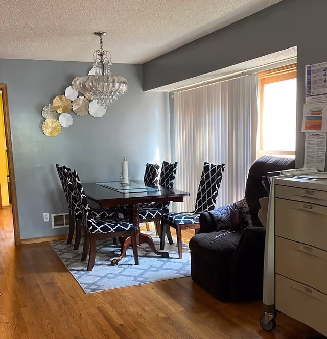 Dining room with glass table, patterned chairs, chandelier, and recliner near window.