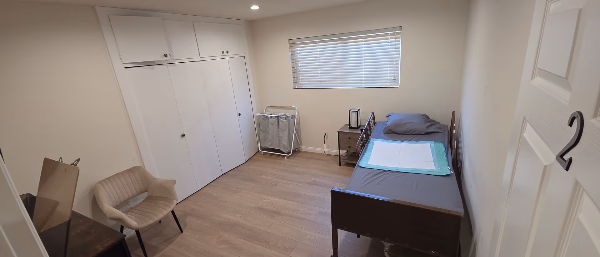 Compact care home bedroom featuring a single bed, wardrobe, chair, and window blinds.