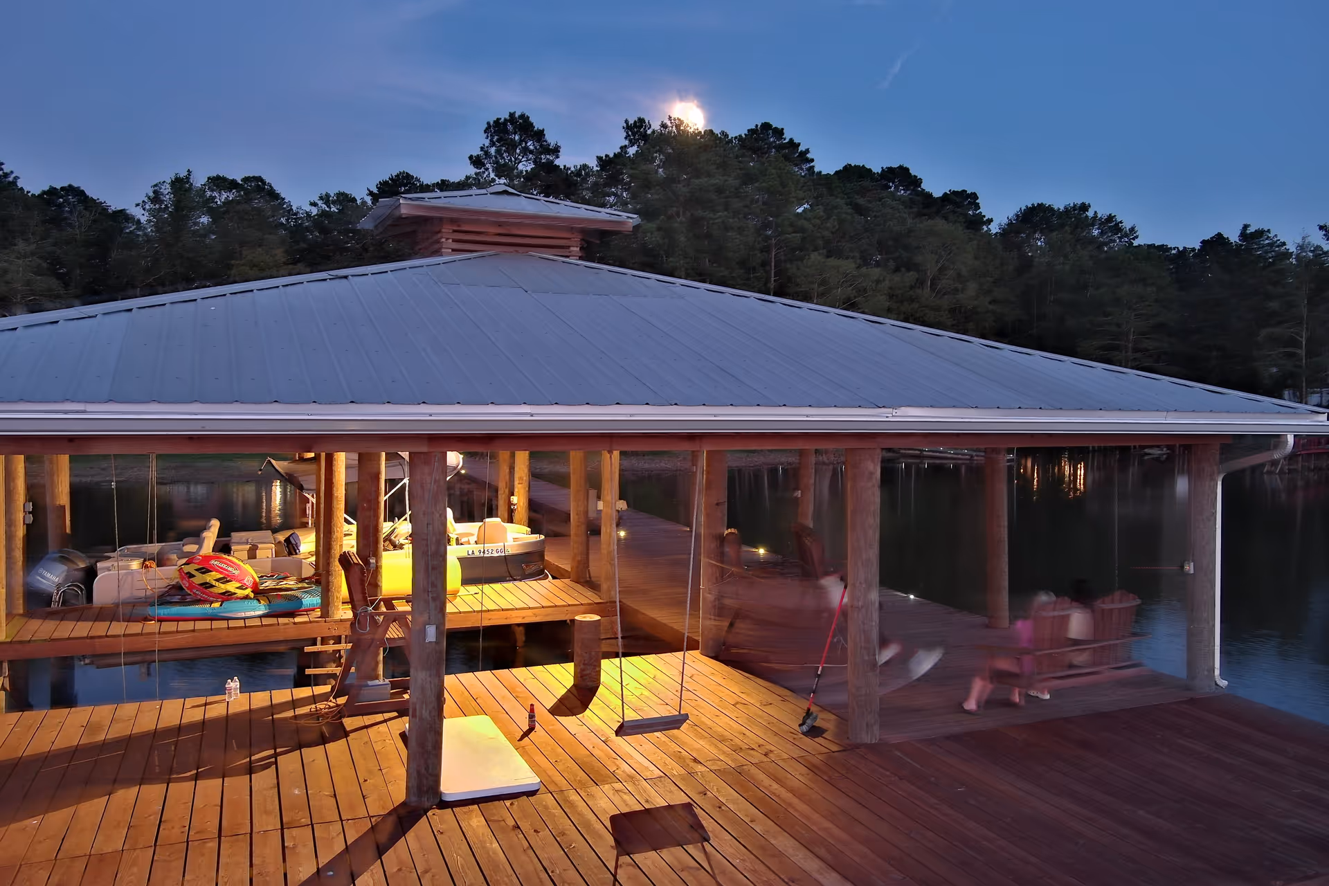 Nighttime view of a covered lakeside dock with boats, swings, and seating.