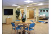 Round table with blue chairs in a bright indoor lounge; wall-mounted TV.
