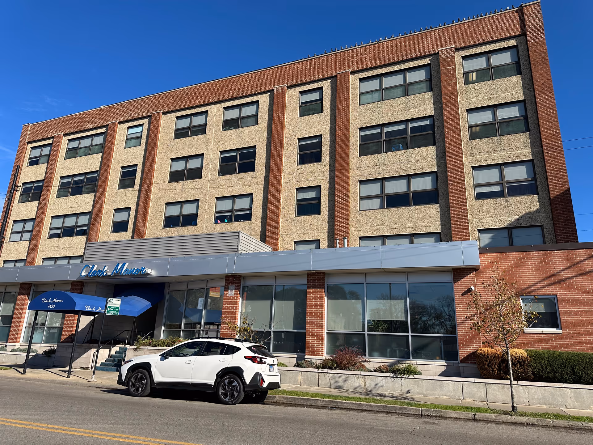 Exterior view of a multi-story brick and concrete building with large windows and a blue awning over the entrance. A white SUV is parked on the street in front of the building. The building has a sign that reads 'Clark Manor'.