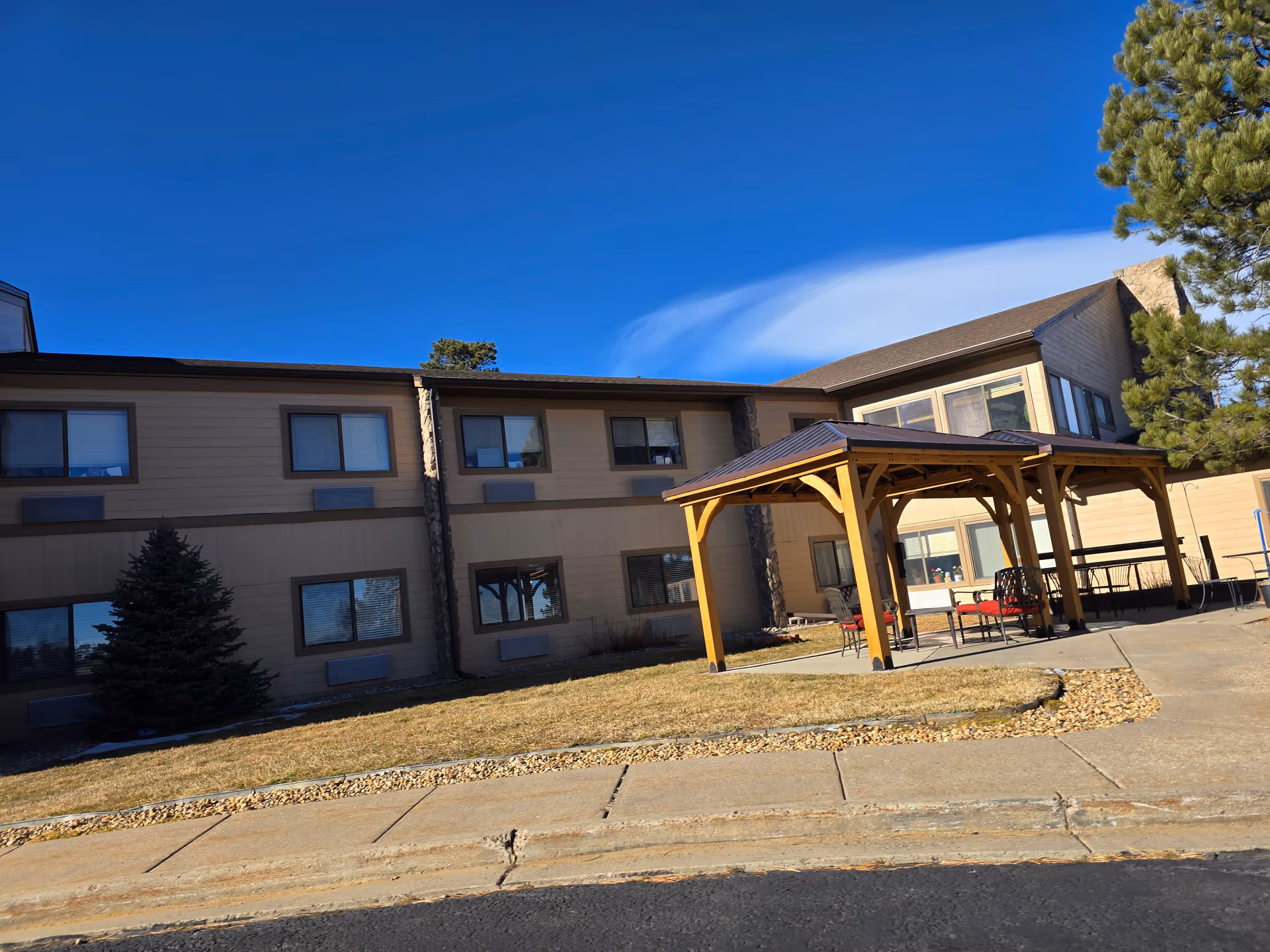 Front exterior of a two-story senior living building with a wooden gazebo and outdoor seating under a clear blue sky.