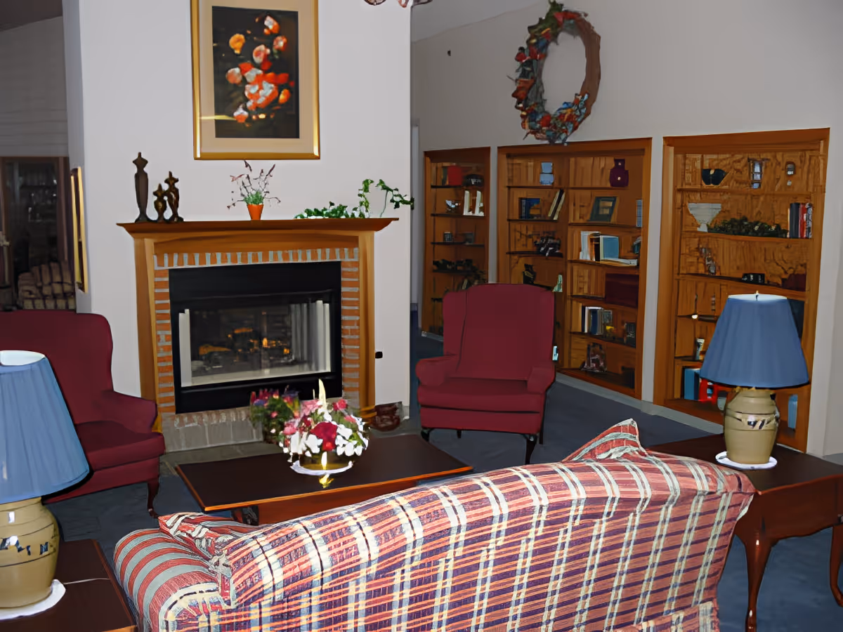 Cozy living room with a plaid sofa, fireplace, armchairs, side lamps, coffee table with flowers, and built-in bookshelves.